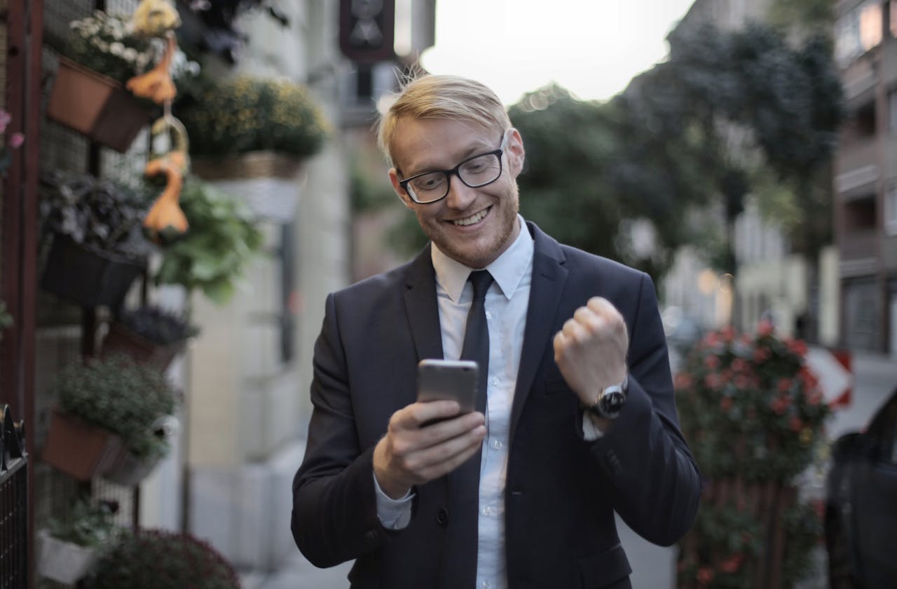 Excited young professional celebrates success outdoors using mobile phone in urban setting.