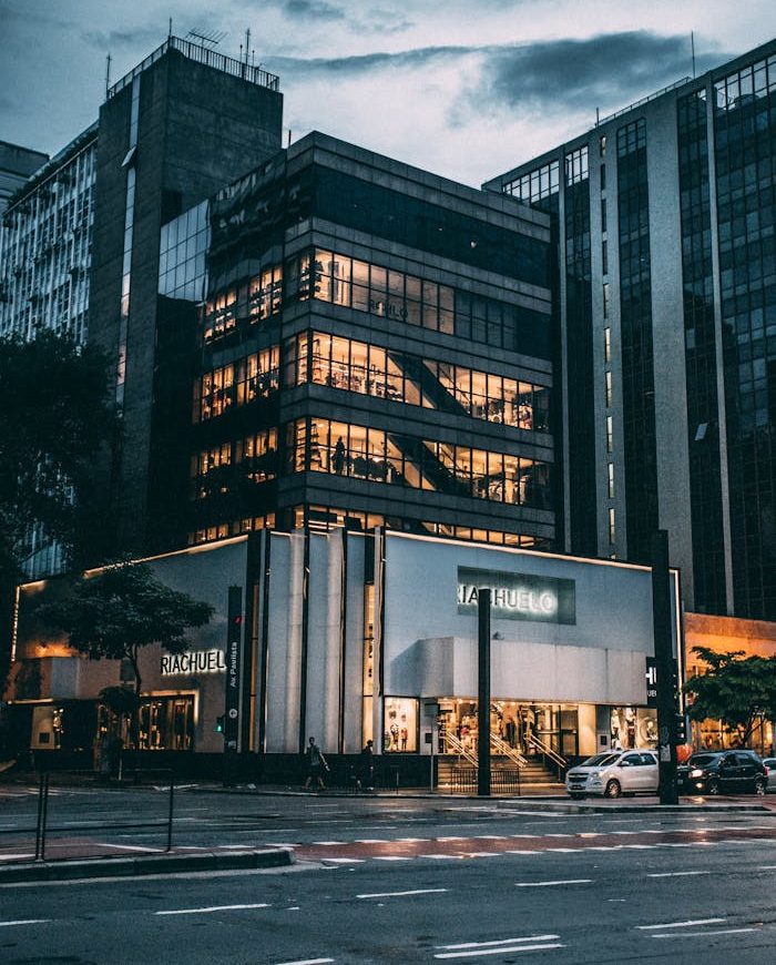 Night view of a city block with illuminated store, modern architecture, and cloudy sky.